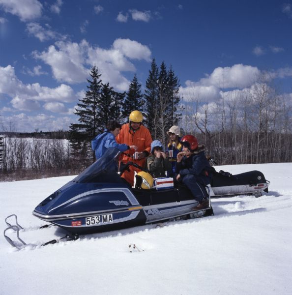 A family of five are outdoors standing around or sitting on their two snowmobiles as they take a break to drink and eat.