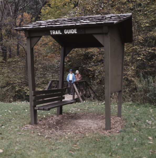 Melvin and Sadie Tvedt of Mount Horeb, walking out of the woods and across a small wooden bridge. In the foreground is a roofed bench with a sign reading: "Trail Guide."