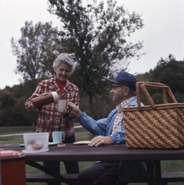 Sadie Tvedt of Mount Horeb, standing and pouring from a thermos liquid into her husband Melvin's cup. A plastic container of food and a picnic basket are sitting on the picnic table. 