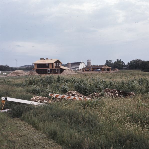 View across field towards a house in the midst of construction. There are barriers near piles of dirt in the foreground, and there are piles of dirt and rock around the house and garage. In the far background is a white barn and two silos.