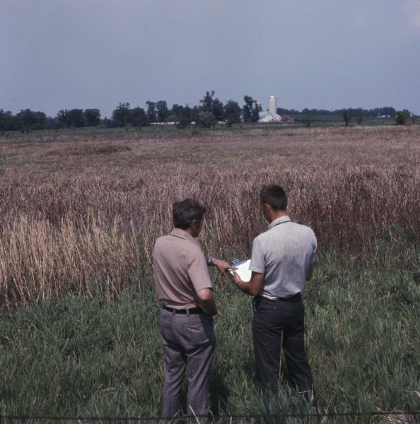 Two men are surveying dried out wetlands. A farm is in the distance.