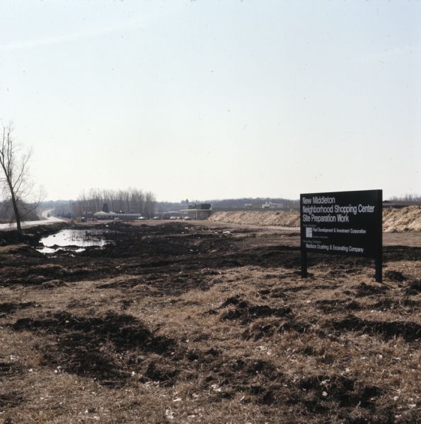 View of a dredged wetland area near a road. A sign along the right reads: "New Middleton Neighborhood Shopping Center Site Preparation Work."