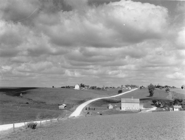 Cumulus clouds are casting their shadows over rolling farmland. Several farmsteads are along a narrow road. There are cows grazing in a pasture. On the reverse of the print is written: "Across the ridges in the dairy region of Wisconsin."
