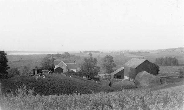 Elevated view from hill of chickens ranging near their coop behind a farmhouse and outbuildings on a tidy farm. There is a large barn on the right with two haystacks behind it. The expansive view beyond the farm includes the southern end of the Horicon Marsh.