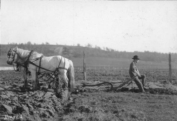 A farmer resting, sitting on the handles of his plow, with his right foot in the furrow. His team, in harness, are standing head to head. They are in front of a barbed wire fence, and a hill is in the background.