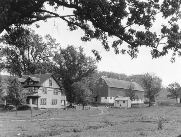 Large branches of an oak tree frame the view of a large farmhouse, barn, and outbuildings. The two-story house has two front dormers and porches on the front and back. A pickup truck is parked behind the house. The barn has a gambrel roof and three roof venilators. Painted on the gable end of the barn is "J.(?). Elmer" with the unreadable date below. There are chickens near the outbuildings. On the reverse of the print is written: "Farm home and buildings in the dairy region of southern Wisconsin."