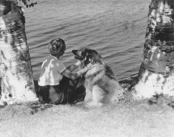 The caption on the reverse of this print reads: "Miss Ferguson sitting with dog on shore of Pine Lake." They are framed by the trunks of two birch trees, and are facing each other with their backs to the camera. The woman is wearing slacks, a blouse, and a paisley patterned bandana. The dog appears to be a collie.