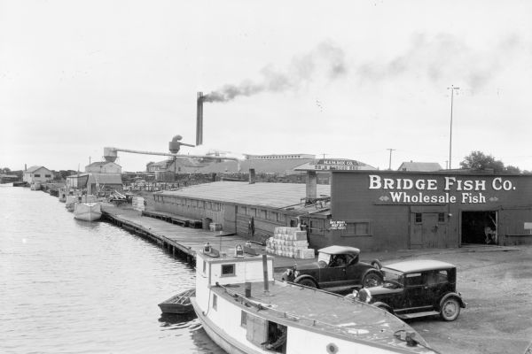 Elevated view of fishing tugs moored alongside the wharf at Marinette. Two automobiles are parked in the foreground, just in front of the Bridge Fish Co. building. Pulp wood is stacked near the buildings of the M. & M. Box Co. in the background. A tall chimney at the rear of the complex is emitting dark smoke.