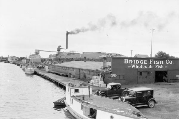 Elevated view of fishing tugs moored alongside the wharf at Marinette. Two automobiles are parked in the foreground, just in front of the Bridge Fish Co. building. Pulp wood is stacked near the buildings of the M. & M. Box Co. in the background. A tall chimney at the rear of the complex is emitting dark smoke. 