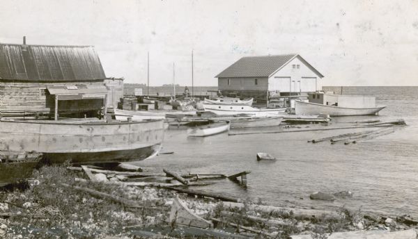 View from shoreline towards fishing boats of various sizes, and sailboats docked at the wharf at Fish Creek. Two persons are carrying gear toward the pier. There is a boathouse at the end of the pier and a small warehouse on the left. A sign on the pier reads: "Thorp Hotel Boat Livery." 