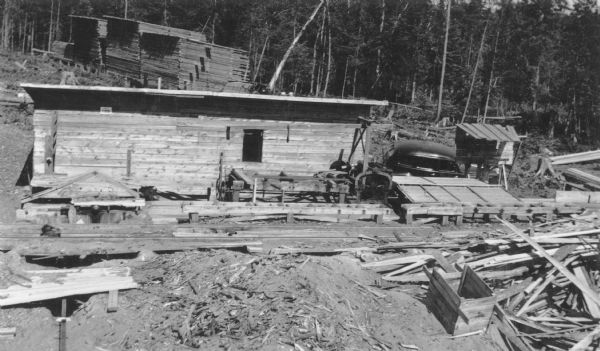 Lumber is stacked in three large piles behind a small wooden shed. There is machinery in front of the shed on the right, and what appears to be a saw pit on the left, with metal tracks crossing the foreground. An automobile is parked to the right of the shed.
