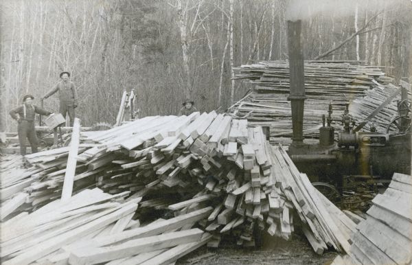 Two men, left, are standing near the circular blade of a sawmill which is set up in a forested area. A third man in the center is peering over a pile of lumber of various dimensions. The steam engine which powered the mill is on the right.  