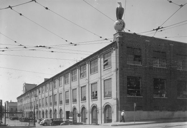 "Old Abe," the bald eagle trademark of the J.I. Case Company, is shown roosting on a world globe with "CASE" written on it, on the corner of a parapet on the front of a three story brick factory building. A man is walking on the sidewalk near the building and automobiles are parked behind a gate and fence along the left side. Overhead are street car power lines anchored to the building.