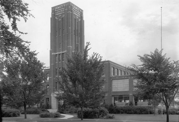 View across street towards the three-story brick factory building, with a taller, square central tower that has symmetrical two-story wings. Identified on the tower as "Amity Leather Products Co.", the Art Deco style building was built in 1924. It housed the Amity Company until 1996, when production was moved to Brown Deer, Wisconsin, and the company name changed to AR Accessories Group.
