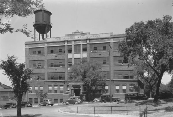 Cars are parked on the street in front of the 1915 brick Parker Pen factory and office building. There is a large water tower on the left. The building was erected in 1915.