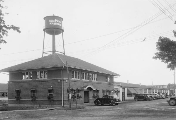 View across street towards automobiles parked in front of a two-story brick office building with a long, one story factory wing. There is a water tower behind the building with "Speed Queen Washers" painted on it. Plants spill from window boxes on the first floor windows, which also have awnings.