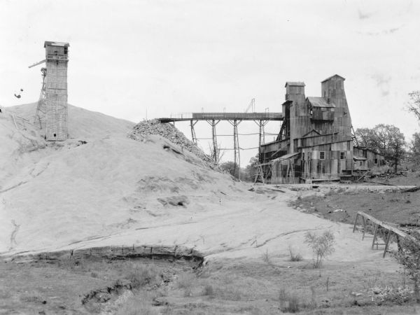 A view of the tailings pile, on the left, and on the right, the shaft house of an abandoned mine. The shaft house has been sided with corrugated metal panels. On the reverse of the print is written: "An old zinc mine in southwestern Wisconsin."
