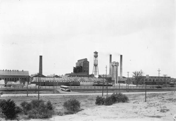 View across field towards a bus sitting in the driveway in front of a large factory complex, which has a water tower and three smokestacks. "Airport 2 Mi." is painted on the roof of the low building in the foreground. The water tower identifies the "Marathon Paper Mills Co."