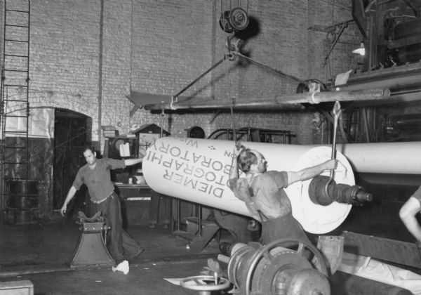 Two men are moving a large roll of paper with the help of an overhead pulley.  