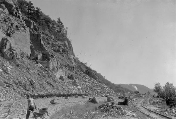 A worker is pulling a wheelbarrow behind him while walking alongside a narrow gauge railroad track at the south end of the East Bluff near Devil's Lake. Empty cars are sitting on the track in the background at the base of a an exposed rocky bluff. There is another set of tracks on the right. A train engine is in the distance. There are other workers in the background.