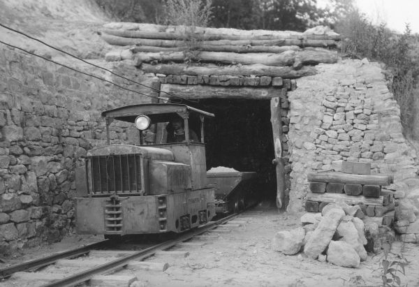 A worker is driving a Plymouth "dinky" locomotive out of the entrance of a sand mine. One full car is behind the engine.  