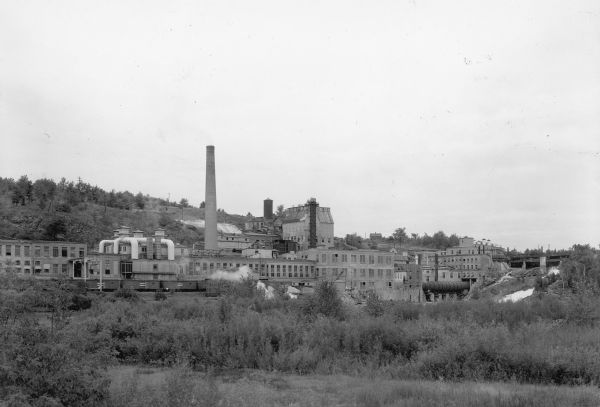 View across field towards large smokestack towering above the complex of buildings at the Niagara paper mill. On the far right, water is flowing through the gates of the dam on the Menominee River.