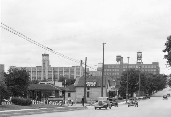 View down street towards two buildings in the background that are part of the Aluminum Goods Manufacturing Company, makers of Mirro cookware. The buildings are identified by signage on two rooftop water towers on the building on the right. The building on the left features a central clock tower. In the foreground is a service station with a long overhang and gas pumps with crown tops. A Coca-Cola sign identifies the business as Vandersteen's Standard Service. There are two boys near the gas pumps, and an older man in a suit is standing on the sidewalk.  