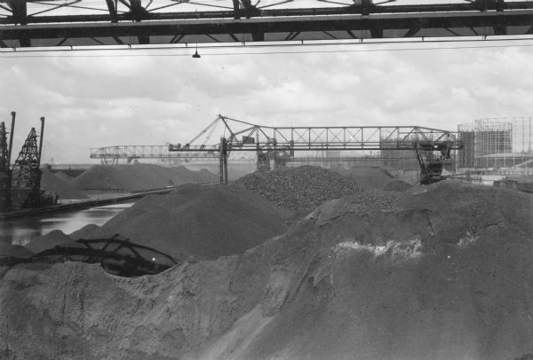 Elevated view of vast piles of coal dominate this scene of the Milwaukee Harbor. A bridge crane is in the center, with two other cranes at the far left. There are three large petroleum storage tanks in the background on the right.