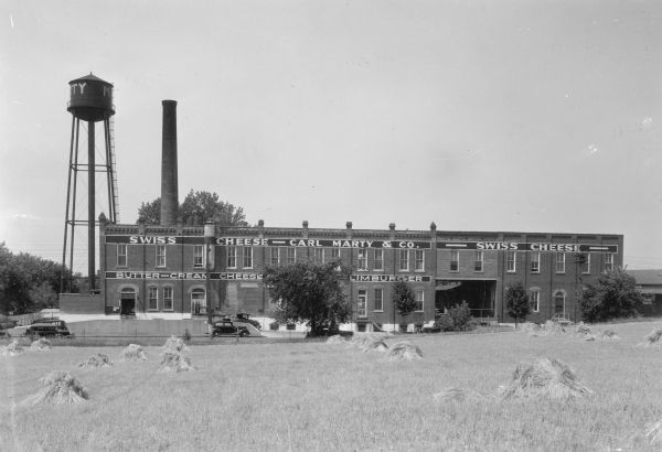 View across harvested field shocks of grain towards the long, two-story brick Carl Marty & Co. factory, featuring a large smokestack and a water tower. There are cars parked in a lot on the left. Painted signage on the building advertises Swiss cheese, butter, cream cheese and limburger. On the reverse of the print is written: "Largest Swiss Cheese Manufacturing Company in the world — Monroe."