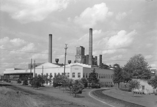 Two large smokestacks rise from the 1903 building of the Rhinelander Paper Company. Railroad tracks are crossing the left foreground diagonally; a second set of tracks are curving off to the right. A sign on the roof at the right side of the building reads: "Rhinelander Paper Co. Manufacturors of Glassine and Greaseproof Papers." There are rail cars at the loading docks on the left.