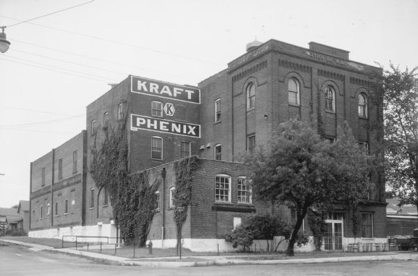 Vines are growing on the side of the brick Kraft Phenix cheese factory building at the corner of Ninth Avenue and Edison Street. Raised letters on the frieze above the front door read: "Antigo Brewing." There are milk cans near the front door, and a pickup truck is parked on the far right. A street light is hanging over the intersection. The Kraft Phenix plant, established by William Kraft in 1924, was the second Kraft cheese manufacturing plant.