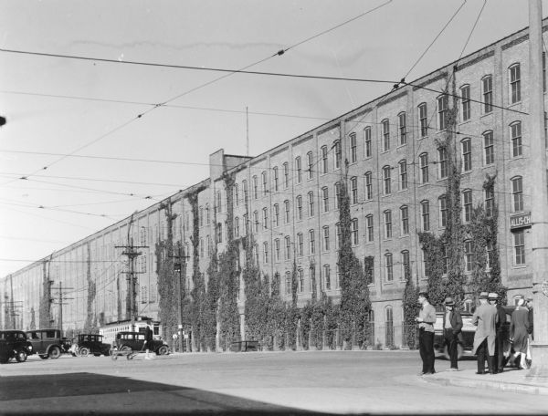 Vines are growing on the facade of the Allis Chalmers factory which extends the full width of this view. There are automobiles and a street car in the background, where a woman wearing high heels pulls a coaster wagon. There are men and a woman near a lamp post in the foreground.  On the reverse of the print is written, "One of the world's largest makers of machines that can't be made — Allis Chalmers."
