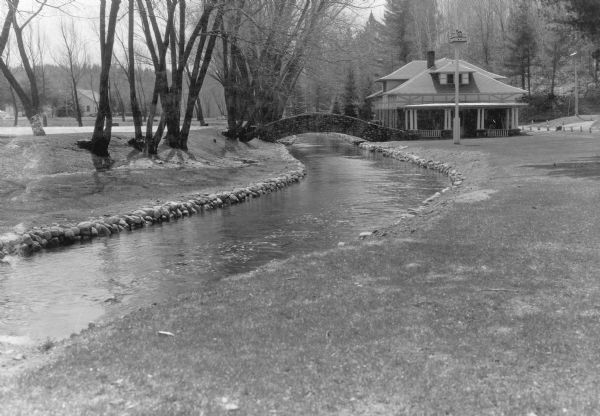 View down a fieldstone-lined stream towards a graceful stone bridge crossing the stream at the fish hatchery. A one and one half story building with a porch, dormers and deep roof overhang is to the right of the bridge. Two bird houses are on tall posts in the yard. On the reverse of the print is written: "Improvement work at Woodruff state fish hatchery (WPA project)."