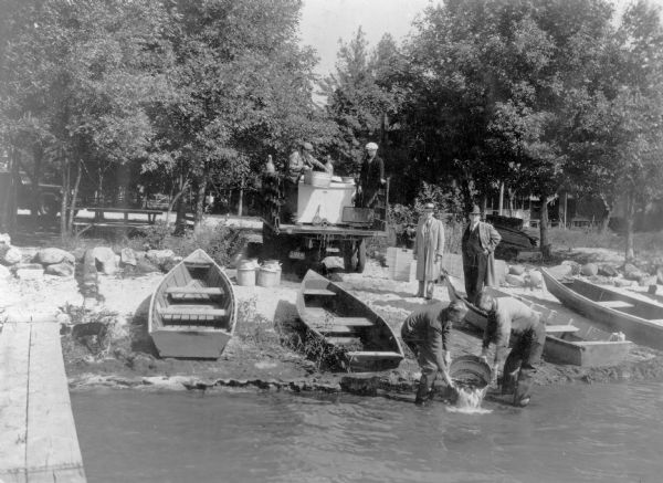 View from pier towards a truck with dual rear wheels parked with the open bed facing the shore of Lake Koshkonong. Two men wearing waders are dumping a basin of young white bass into the lake as two other men are watching from the shore. There are two men standing in the bed of the truck which is holding two oxygen canisters and a large rectangular vat. Rowboats have been pulled up onto the shore. The description on the reverse of the print reads: "One of the Conservation Department's new oxygen tank cars in the state fish distribution service delivering white bass to Lake Koshkonong. Showing Howard Berquist, operator, Tony Niber, Assemblyman Palmer Daugs and Warden William P. Elliott. Nelson Hake and C.L. Sullivan are putting fish in the lake."