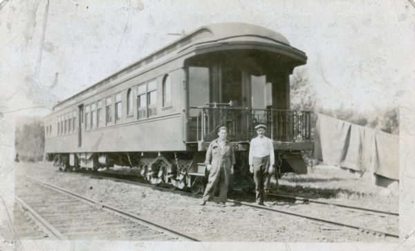 Two men are standing in front of a railroad car on railroad tracks. A blanket is hanging over a line tied to the railing of the car. On the reverse of the print is written: "Frank Suthers and assistant with the old Conservaton department fish car. This car was purchased as a bargain in 1912 and outfitted as living quarters for Conservation Dept. personnel and for transportation of young fish for stocking waters throughout the state."