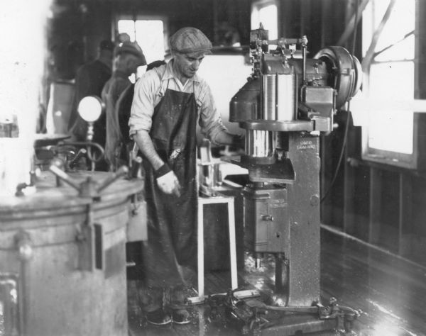 A man wearing a long leather apron, cap and gloves is operating a machine that secures lids to cans. Written on the reverse of the print is the description: "Canning carp. Capping cans to be placed in cookers." There is a pressure cooker in the foreground on the left.