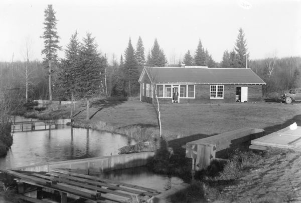 Slightly elevated view towards an unidentified man standing in the doorway of a simple wood frame, single story building. A sign on the roof identifies the "State Fish Hatchery." In the foreground, barriers define pools while allowing stream water to flow through. There is an automobile parked on the far right.