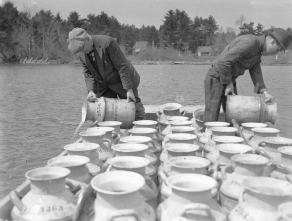 Two unidentified men, working from a boat or raft, are dumping the contents of milk cans into the waters of a lake. There are small cottages on the shore in the background. On the reverse of the print is written: "Planting pike. Lake Vieux Desert." 