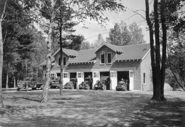 A one and one half story masonry block building with five bays with open doors stands in a wooded area.  A track vehicle on a trailer is hitched behind a large truck at far left; another track vehicle is visible in the bay at far right.  Three small trucks occupy the center bays. The written caption on the reverse identifies the "New forest protection division garage at Rhinelander ranger station, with fire equipment ready for call."