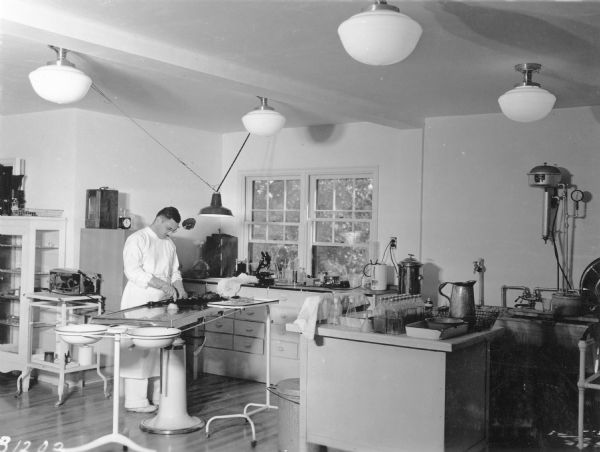 A man wearing a white surgical gown and gloves is standing at a surgical table. He is performing a necropsy on a small animal. There is a double basin standing at the foot of the table. On the far left is a portable sterilizer. At right is a work area with utility sinks, bottles and flasks as well as enameled metal trays.