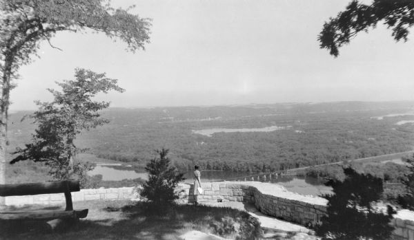View looking down towards a woman wearing a striped dress and holding a purse standing at a low stone wall at the Point Lookout observation area above the Wisconsin River, just upstream from where it joins the Mississippi River. Several sloughs as well as a railroad bridge are in the far background. Trees in the foreground frame the view. There is a rustic log bench on the left.