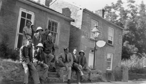 A group of eight young men, all Georgia Junior Rangers, and two of their guides, are posing in front of Pendarvis House, a Cornish miner's cottage. Five men are standing on the lawn, while the other men are sitting on the stone wall in front of the cottage. Three of the junior rangers are wearing pith helmets. Trelawny House is on the right.  Restoration of the two houses was nearly complete. The two upright stone slabs in front of Trelawny are supports for a gate in the later-constructed stone wall. 
