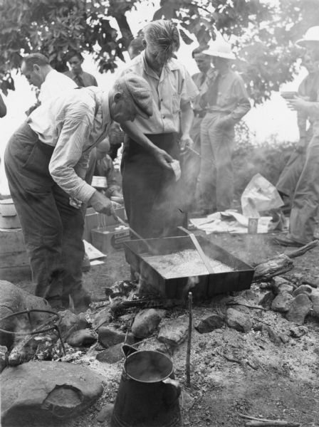 Joe Norton, left, and his son Bud are tending a large pan of fish chowder cooking over the coals of a campfire on the shore of Green Lake.  In the background are several Georgia Junior Rangers and their tour guides. Two of the rangers are wearing pith helmets.