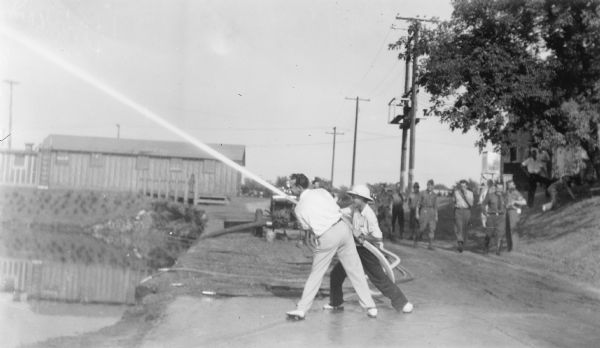 Two men, one wearing a pith helmet, are directing the stream from a fire hose over a body of water. There are several men in the background, some wearing Wisconsin Conservation Department uniforms. There are utility poles and a low building in the background. The description on the reverse of the photograph reads: "Eugene Able & Jerome Webb try their skill at fire fighting at the forest protection headquarters, Tomahawk."