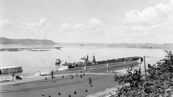 Elevated view of a coal barge pushed by a stern wheeled steamboat heading upstream from the Mississippi River Lock and Dam No. 9 north of Prairie du Chien, entering Lake Winneshiek. There is a small tug nearby. A car is parked near the lock and there are two people standing on the shore. Young trees are growing in a grassy area. In the foreground are utility lines and railroad tracks.