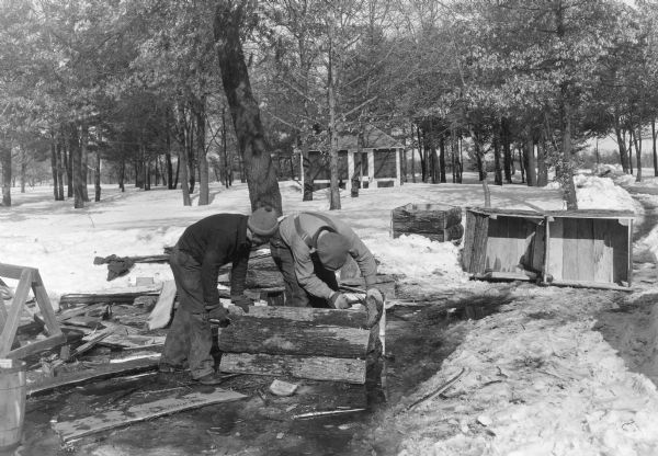 Two men in work clothes are bending over a box they are constructing from slab wood near Lake Menomin. There is snow on the ground and a park shelter in the background. The description on the reverse of the photographs states: "WPA workmen are using lumber obtained from dead or undesirable trees to construct bass spawning boxes on a WPA lake and stream improvement program. These boxes are filled with gravel and sunk in batteries on muddy lake bottoms to provide ideal spawning conditions for bass."