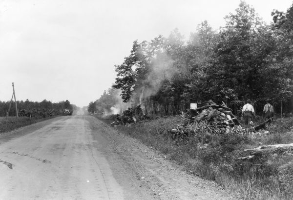 Smoke is rising from piles of burning brush and stumps along a country road. Two men are walking near another pile of brush in the foreground on the right. A sign on that pile reads: "USA WORK WPA." There is a truck parked along the road in the background. A description on the reverse of the photograph states: "Roadside cleanup work carried on by WPA crews in Willard. Clearing away of brush, slash, and other highly inflammable material along roadsides does much to prevent fires from starting and gaining headway." 