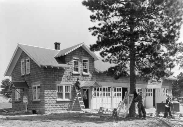 One man is working on a ladder near the side door of a one and one half story, masonry block building with five garage bays. There are two men on the right, with one of the men carrying a shovel, where forms have been built for the driveway. A sign near the front door reads: "USA WORK WPA." A description on the reverse of the photograph reads: "Ranger station and garage built by the WPA at Conover. It is one of 21 similar buildings under construction by the WPA in forest protection districts to provide convenient office headquarters for forest rangers and adequate storage space for tools, trucks, and fire-fighting equipment."