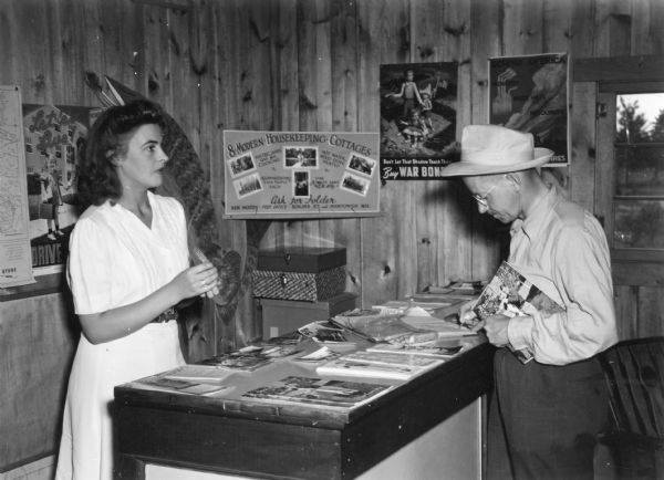 A young woman in a light-colored dress is standing behind a counter as an older man is writing in a notebook. He is holding a tourism brochure for Boulder Junction. There are photographs, maps and other brochures on the counter. Several posters are tacked to the knotty pine paneled walls, including a school zone safety poster, an advertisement for "8 Modern Housekeeping Cottages" in Boulder Junction and Manitowish, and Lawrence Beall Smith's 1942 War Bonds Poster featuring three children in the shadow of a swastika.  