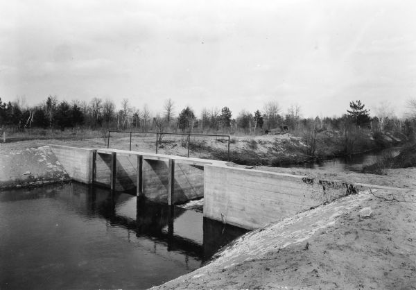 A view looking downstream at a recently built concrete dam with four gates. A note on the reverse of the photograph reads: "Dam built by WPA in drainage ditch. More than 30 dams of this type have been placed in drainage ditches in this Central Wisconsin area to check the uncontrolled drainage which has lowered the ground moisture level until the surrounding country has become virtual desert."